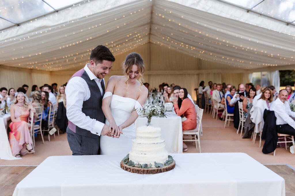 A bride and groom smile as they cut their wedding cake together under a white tent, surrounded by seated guests who are watching and celebrating the moment.