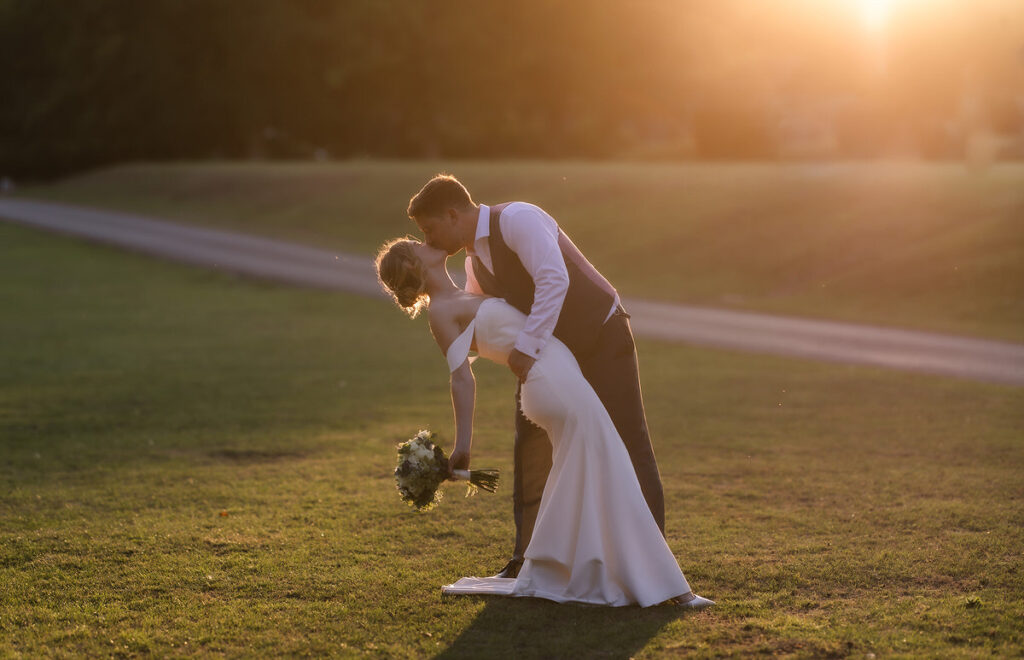 A bride and groom share a romantic dip and kiss on a grassy field at sunset, with the bride holding a bouquet and warm sunlight glowing behind them.