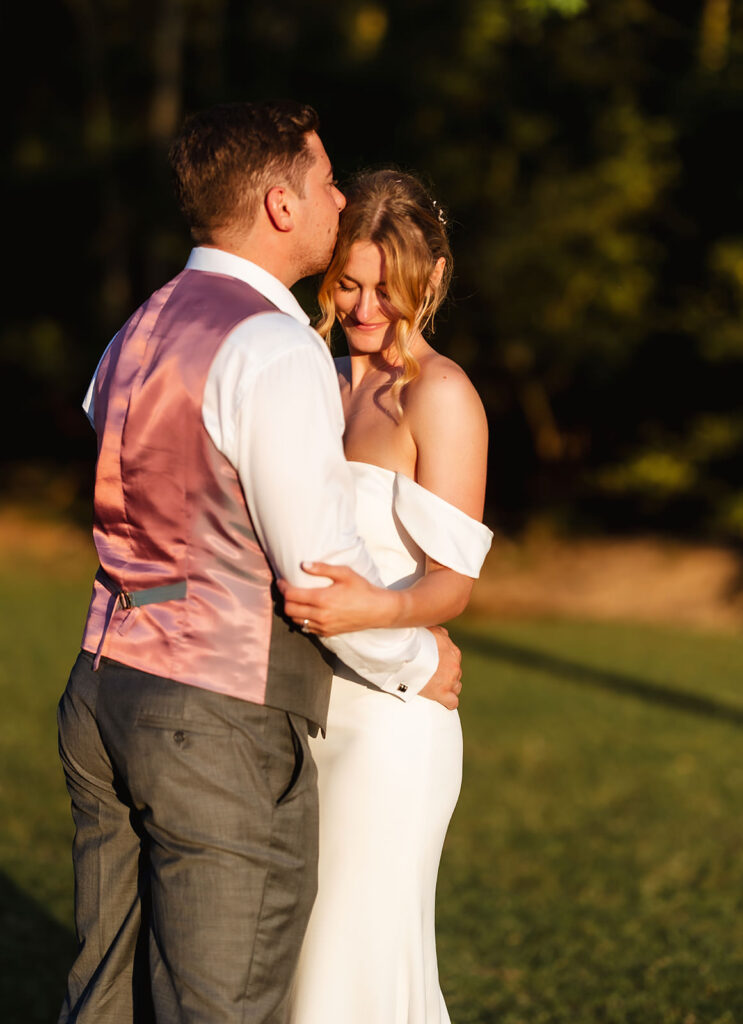 A groom in a pink vest embraces and kisses the forehead of his smiling bride in an off-shoulder white dress outdoors, with sunlight casting a warm glow on the couple.