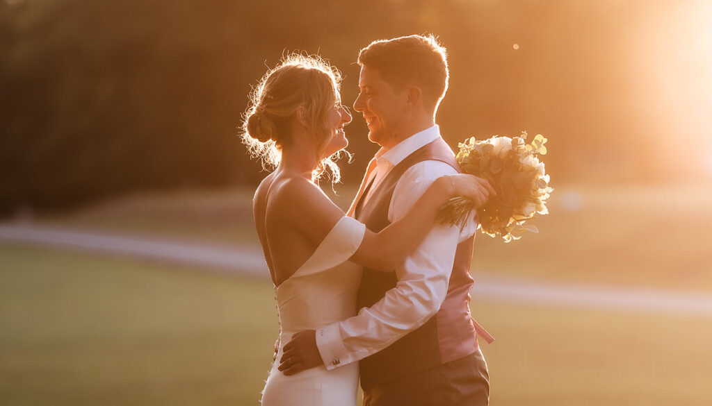 A bride and groom embrace outdoors at sunset, smiling at each other. The bride holds a bouquet of flowers, and warm sunlight creates a romantic, golden glow around them.