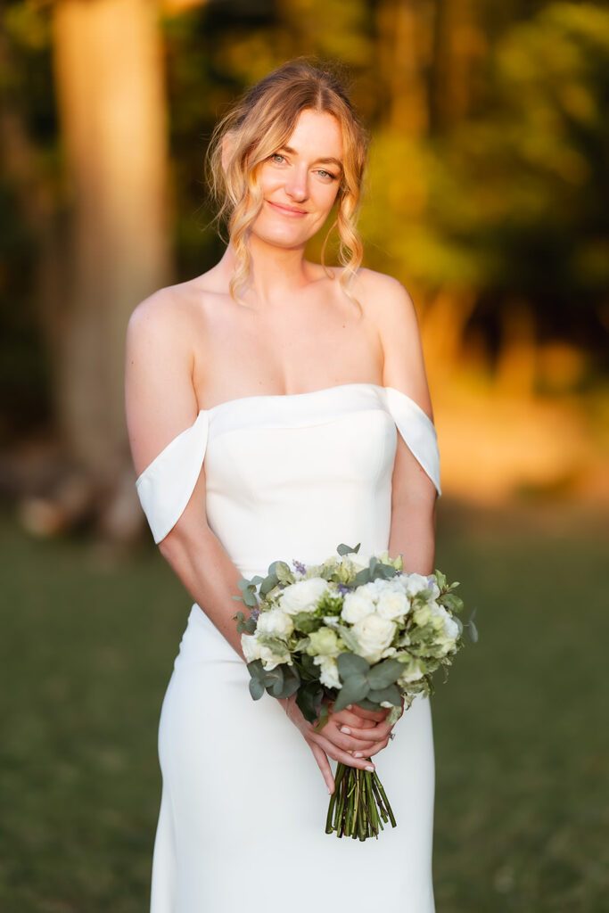 A woman in an off-shoulder white wedding dress stands outdoors, smiling softly and holding a bouquet of white and green flowers, with sunlight and trees in the blurred background.