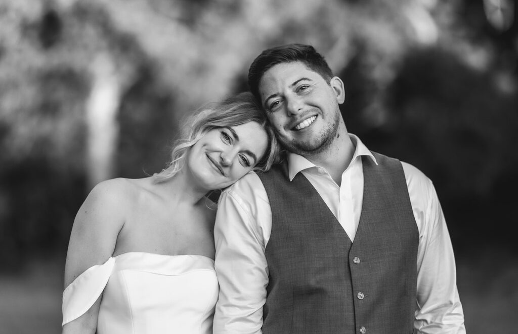 A smiling couple poses outdoors; the woman in a white off-shoulder dress rests her head on the shoulder of the man, who wears a vest over a dress shirt. The background is blurred. Black and white photo.