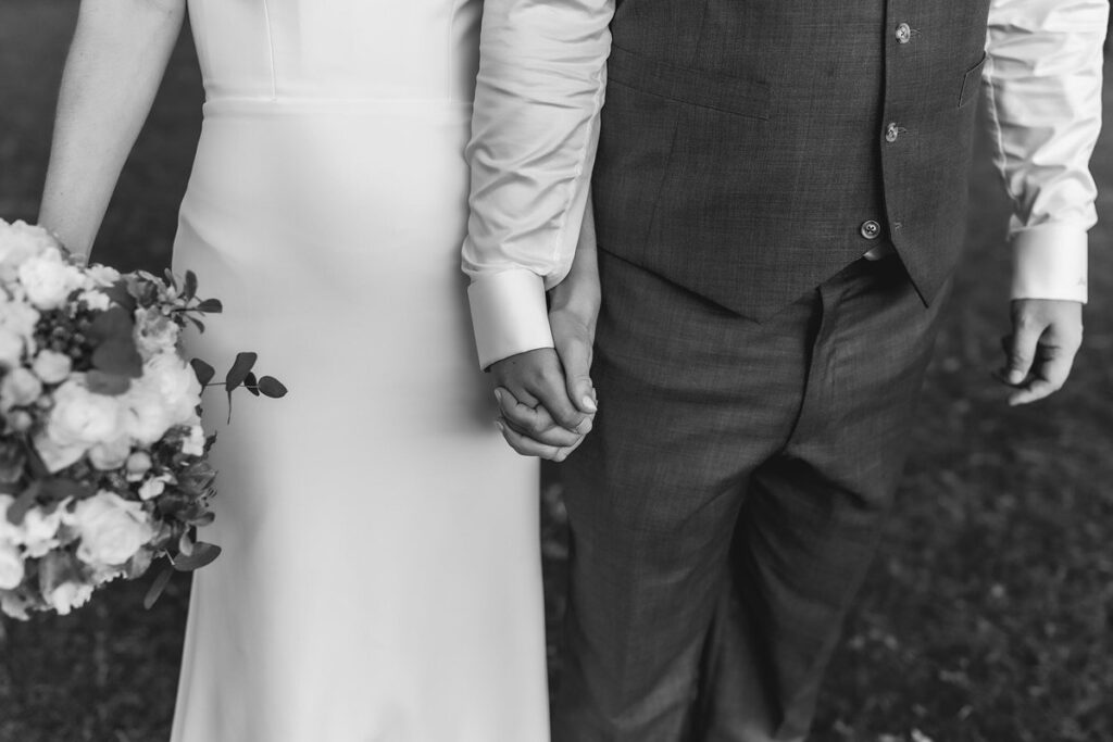 A black and white photo showing a couple holding hands. The woman is wearing a white dress and holding a bouquet, while the man is in a suit. Only their torsos and hands are visible.