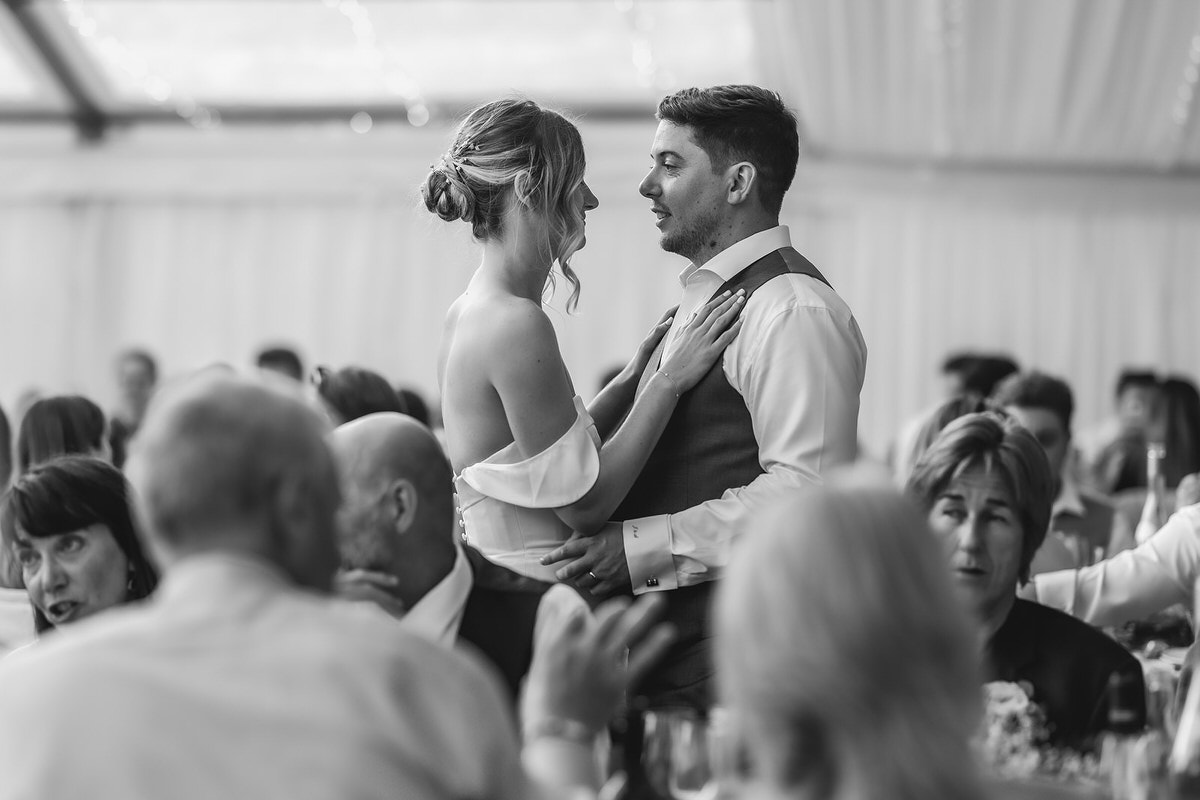 A bride and groom share a close moment on the dance floor at their wedding reception, surrounded by seated guests. The image is in black and white, capturing an intimate and joyful atmosphere.