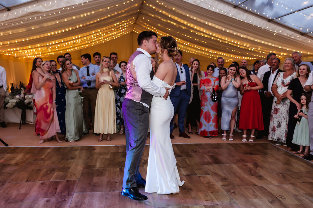 A bride and groom share their first dance in a warmly lit, decorated marquee, surrounded by smiling, applauding guests dressed in colorful attire. String lights hang above, creating a festive and romantic atmosphere.