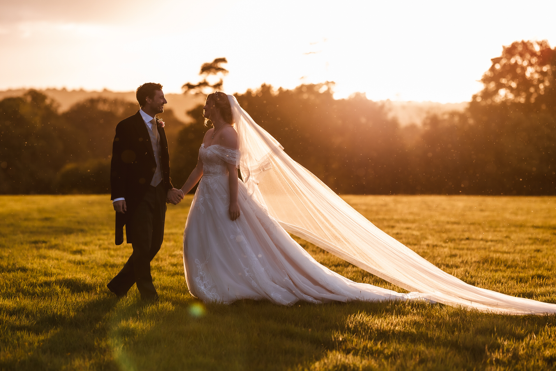 A bride and groom hold hands and walk across a sunlit field at sunset; the bride wears a long white gown with a flowing veil, and the groom is in a dark suit. Captured beautifully by a Hampshire wedding photographer, trees and sunlight fill the background.