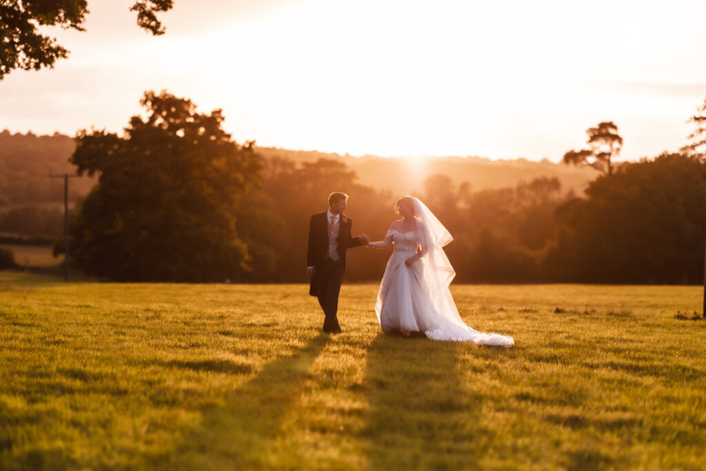 A bride and groom walk hand in hand across a grassy field at sunset, their shadows stretching behind them. Captured by a Hampshire wedding photographer, the golden sunlight and trees create a magical backdrop for this unforgettable moment.