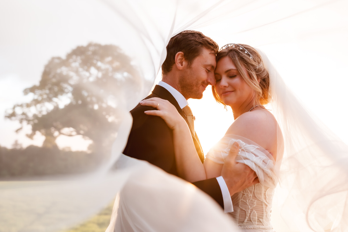 A bride and groom embrace outdoors at sunset, touching foreheads and smiling softly as the bride’s veil gently wraps around them, sunlight filtering through—captured beautifully by a Hampshire wedding photographer.
