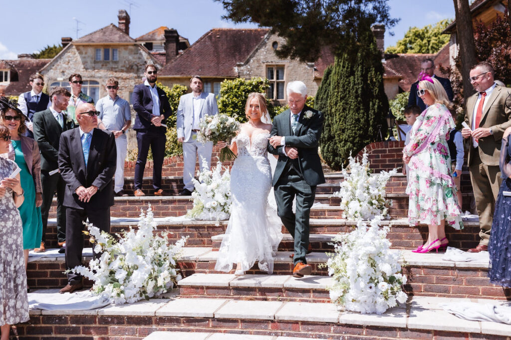 A bride in a white gown walks down outdoor brick steps, holding a bouquet and accompanied by an older man in a suit, surrounded by smiling guests and white floral arrangements on a sunny day.