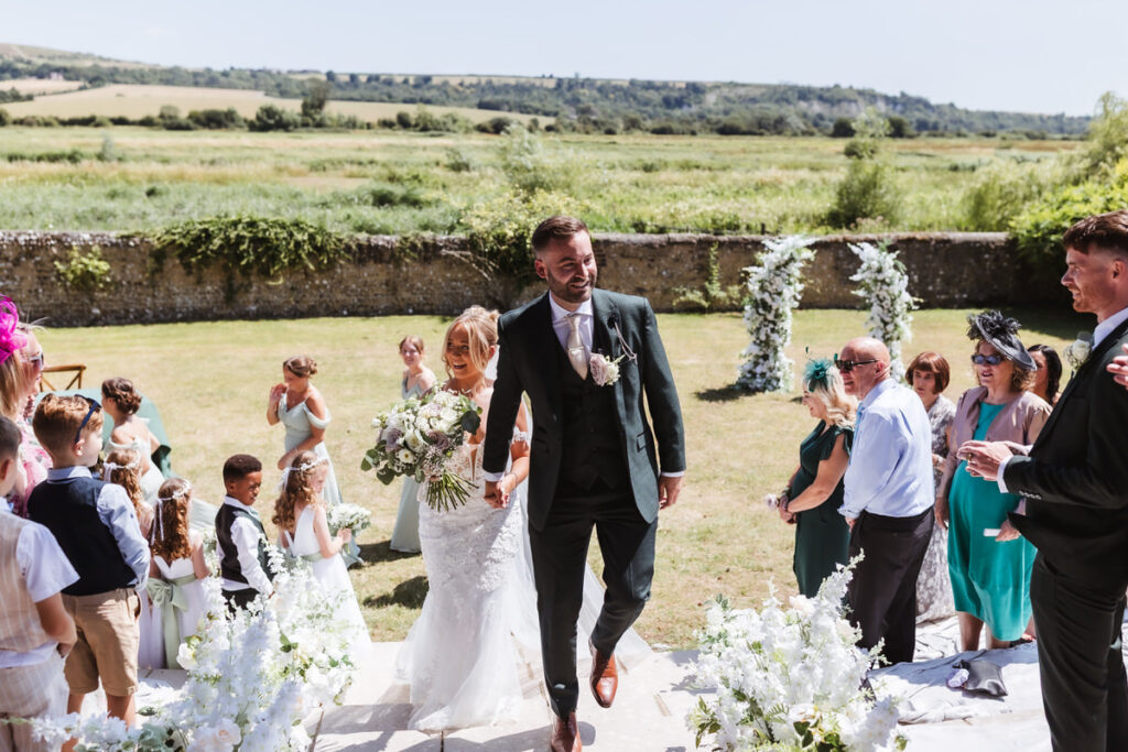 A bride and groom walk up stone steps outdoors, holding hands and smiling, surrounded by guests clapping and celebrating, with lush greenery and hills in the background.