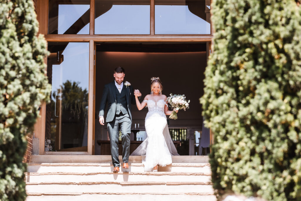A bride in a white dress holding a bouquet and a groom in a dark suit walk hand-in-hand down outdoor steps, with greenery and a building entrance in the background.