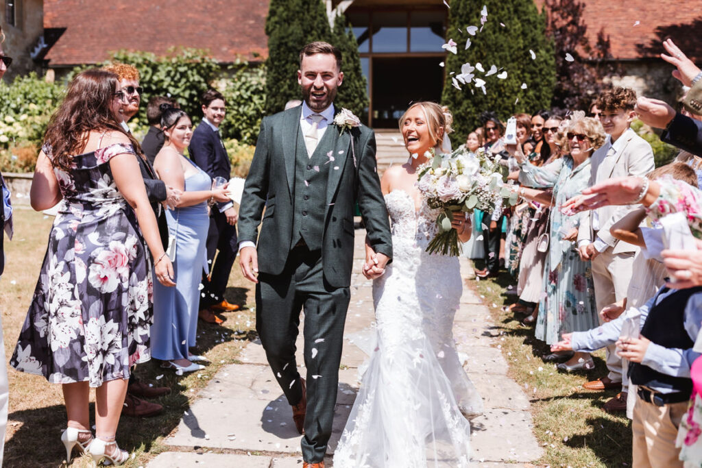 A bride and groom walk hand in hand down an outdoor aisle, smiling as guests on both sides throw flower petals in celebration. The scene is sunny, festive, and surrounded by greenery.