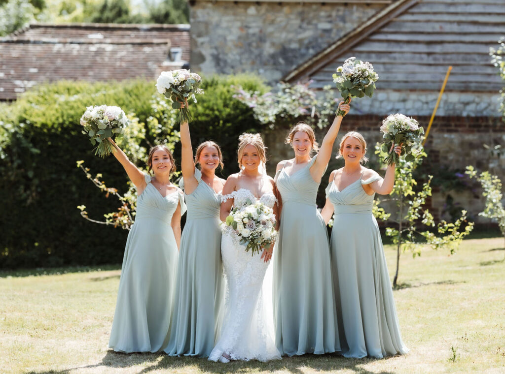 Five women in pale blue dresses, one in a white bridal gown, stand outdoors on grass in front of a rustic building. The four bridesmaids hold bouquets aloft, smiling, with greenery in the background.