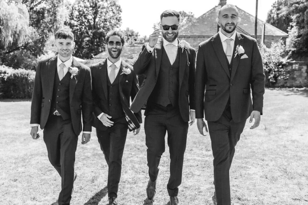 Four men in suits and boutonnieres walk together on grass, smiling and laughing, on a sunny day with trees and a building in the background. The photo is in black and white.