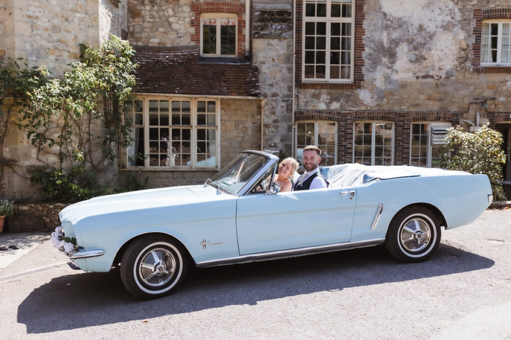 A bride and groom sit smiling in a light blue vintage convertible car parked outside a rustic stone building with large windows and climbing plants.