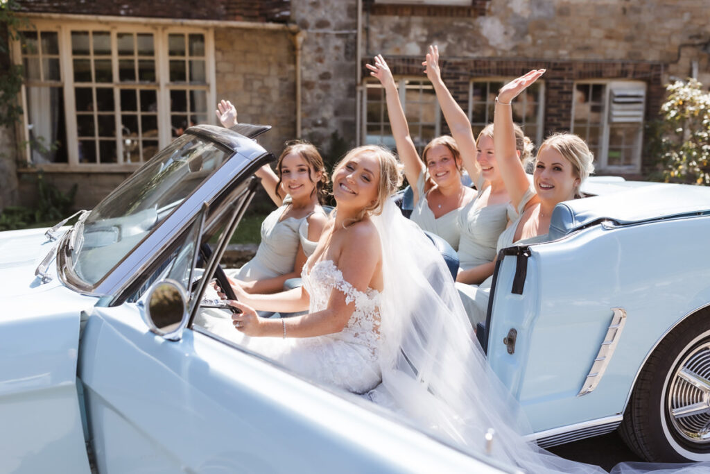 A bride in a white dress and veil sits in the driver’s seat of a light blue convertible with four bridesmaids in pale dresses, all smiling and waving, outside a rustic stone building.