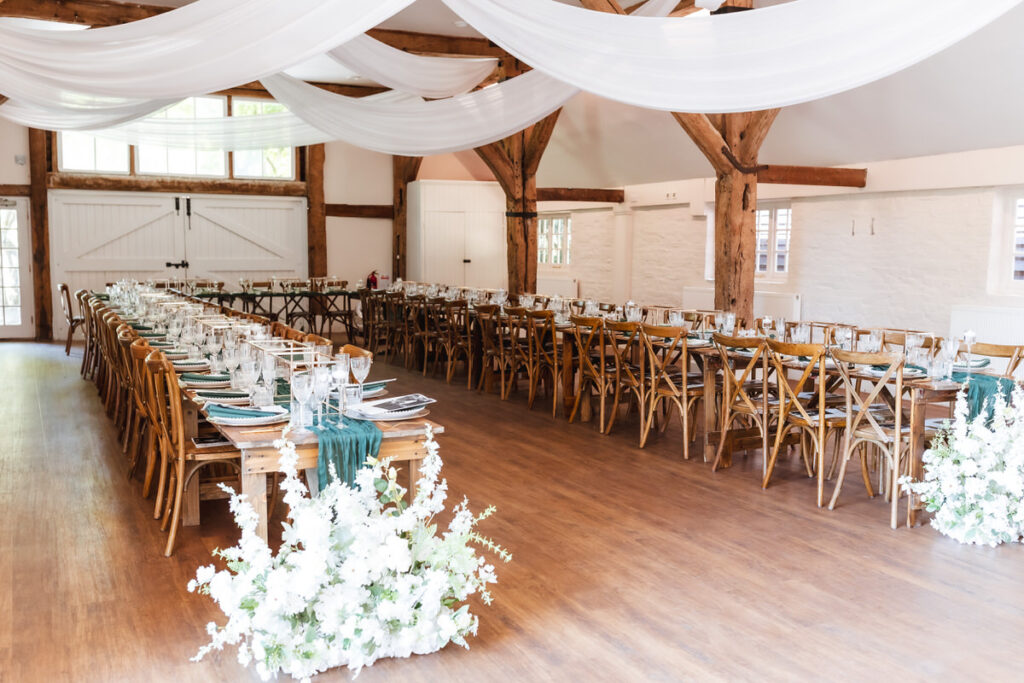 A rustic wedding reception hall with long wooden tables, wooden chairs, white place settings, teal table runners, and white floral arrangements. White drapes hang from the ceiling, and natural light fills the room.