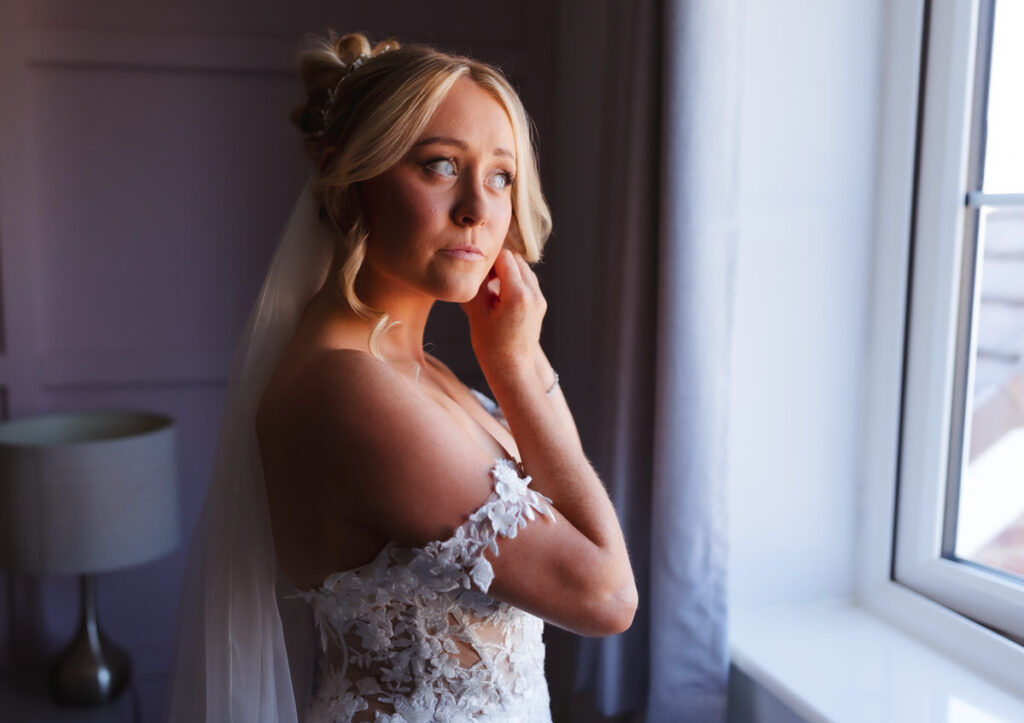 A bride in a white lace wedding dress stands near a window, touching her earring, and gazes outside thoughtfully. Soft natural light illuminates her face and veil.