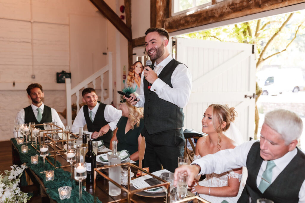 A man in a suit stands giving a speech at a wedding reception, holding a microphone. Other guests, including the bride in a white dress, sit smiling at a long table decorated with candles and greenery.