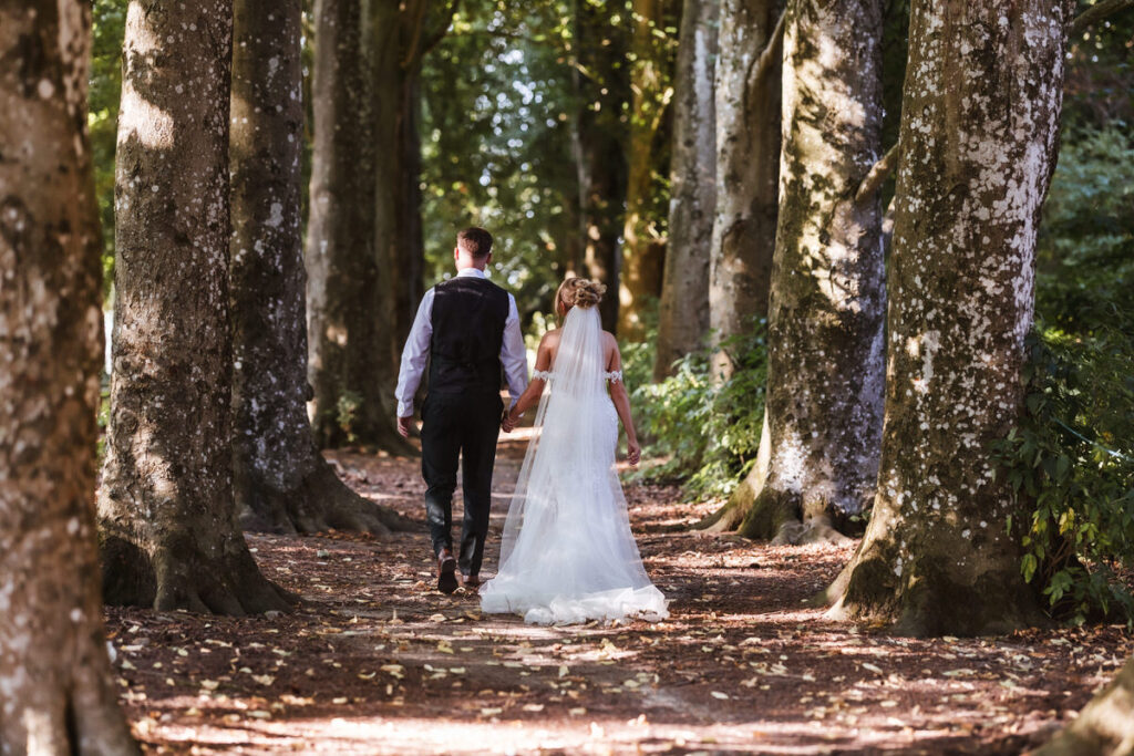 A bride and groom walk hand in hand down a sunlit, tree-lined path, surrounded by tall trees with dappled light filtering through the leaves. The bride wears a white dress and veil; the groom wears a vest and dress shirt.