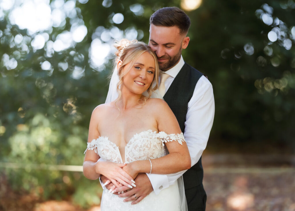 A bride in a white lace dress and a groom in a suit embrace outdoors, smiling and standing close together with trees blurred in the background.
