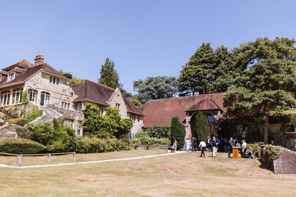 A group of people gather on a sunny lawn outside picturesque stone and brick buildings with sloped red roofs and surrounded by green trees.