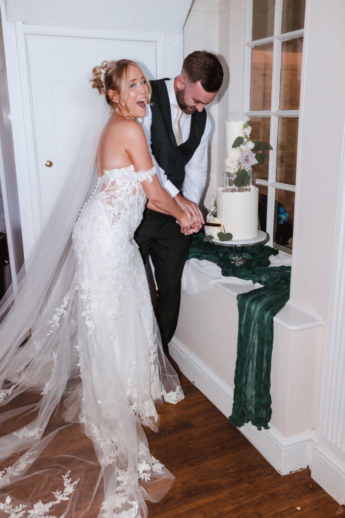 A smiling bride and groom in wedding attire cut a white cake together, standing next to a round table draped with a dark green cloth in a bright room with wooden floors.