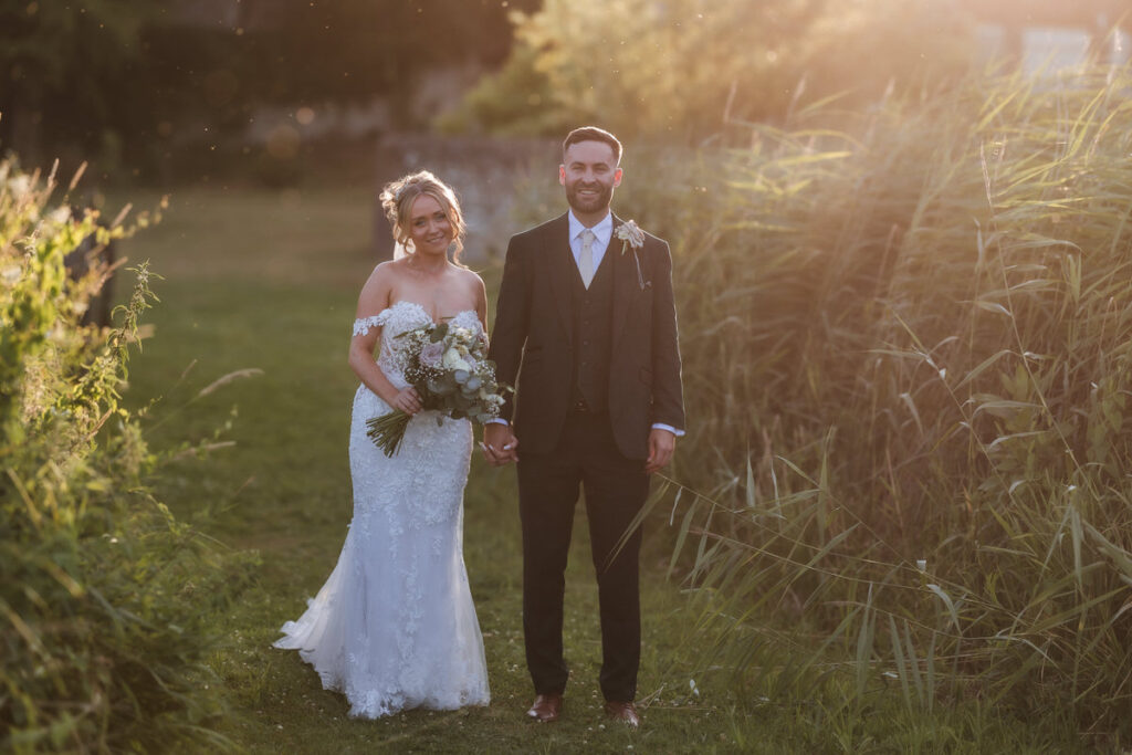 A bride in a white lace gown and a groom in a dark suit stand holding hands outdoors, surrounded by greenery, with warm sunlight shining behind them.