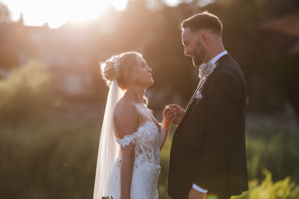 A bride and groom stand outdoors at sunset, holding hands and smiling at each other. The bride wears an off-shoulder lace dress and veil, while the groom is in a suit with a boutonniere. Soft sunlight glows behind them.
