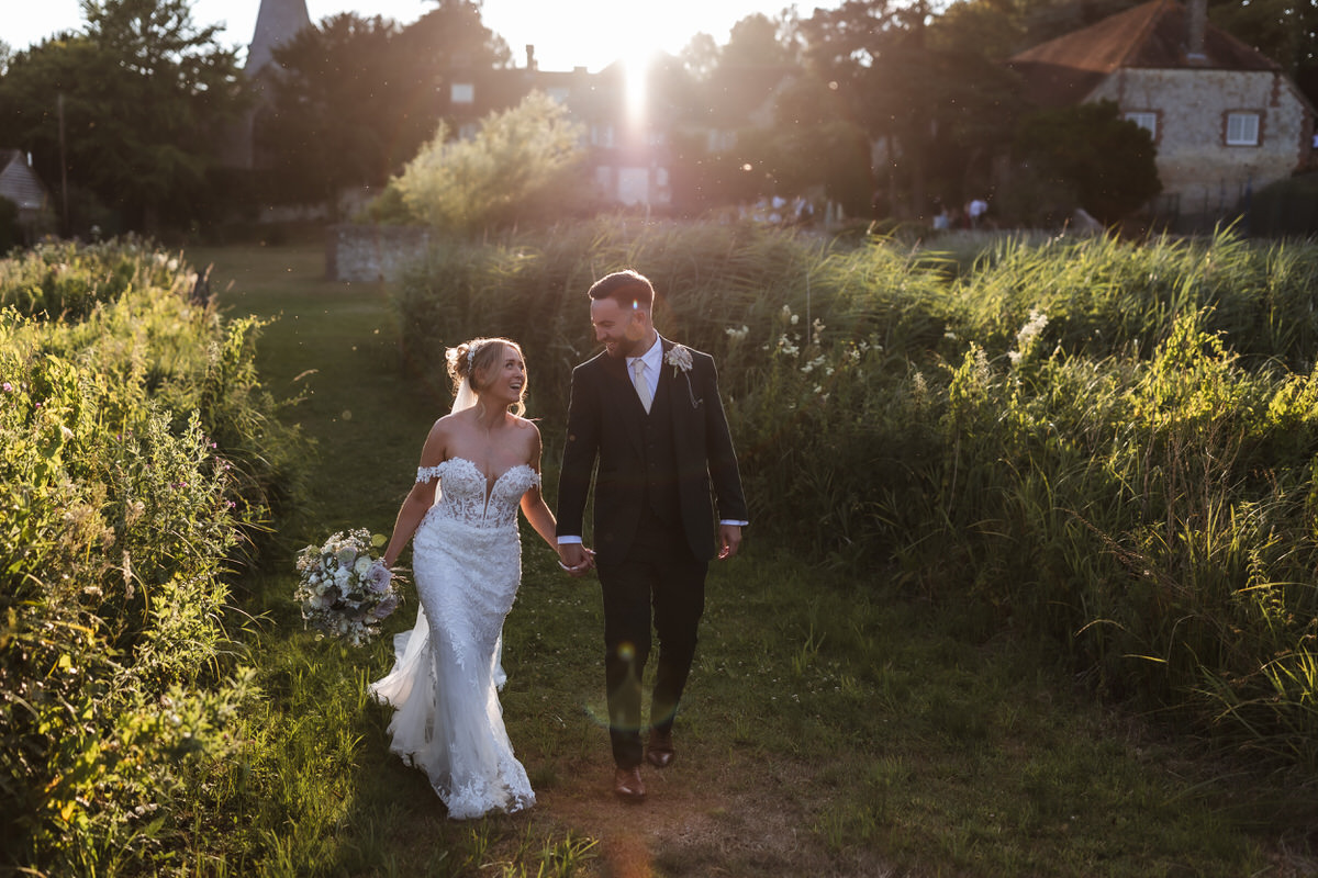 A bride and groom walk hand in hand outdoors at sunset, smiling at each other. The bride wears a white dress and holds a bouquet, while the groom wears a dark suit. They are surrounded by lush greenery and soft sunlight.