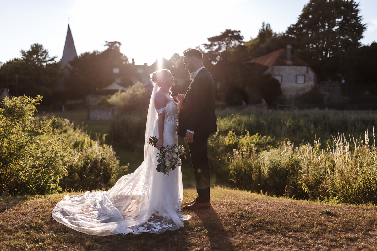 A bride and groom stand facing each other outdoors at sunset, with the bride holding a bouquet. Sunlight shines behind them, creating a romantic, glowing effect. Lush greenery and buildings are visible in the background.