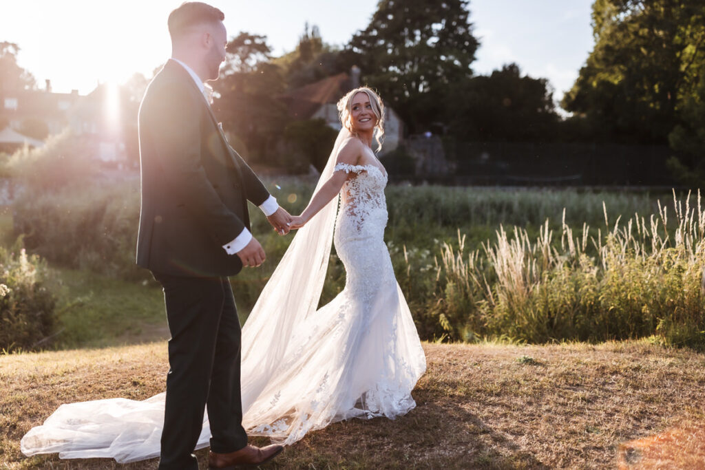 A bride in a white lace gown and a groom in a dark suit hold hands outdoors, smiling at each other, with sunlight shining through trees and greenery in the background.