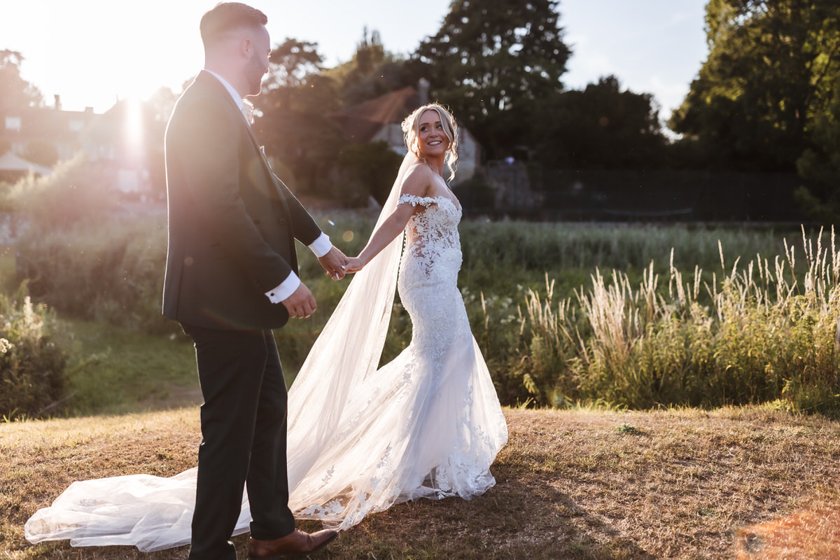 A bride in a lace wedding dress and a groom in a dark suit hold hands and smile at each other while walking outdoors in sunlight, with green grass and trees in the background.