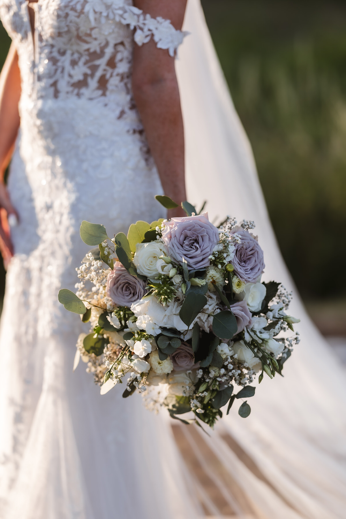 A bride in a lace wedding gown holds a bouquet of pale purple and white roses, babys breath, and greenery, with sunlight highlighting the flowers and her flowing veil.