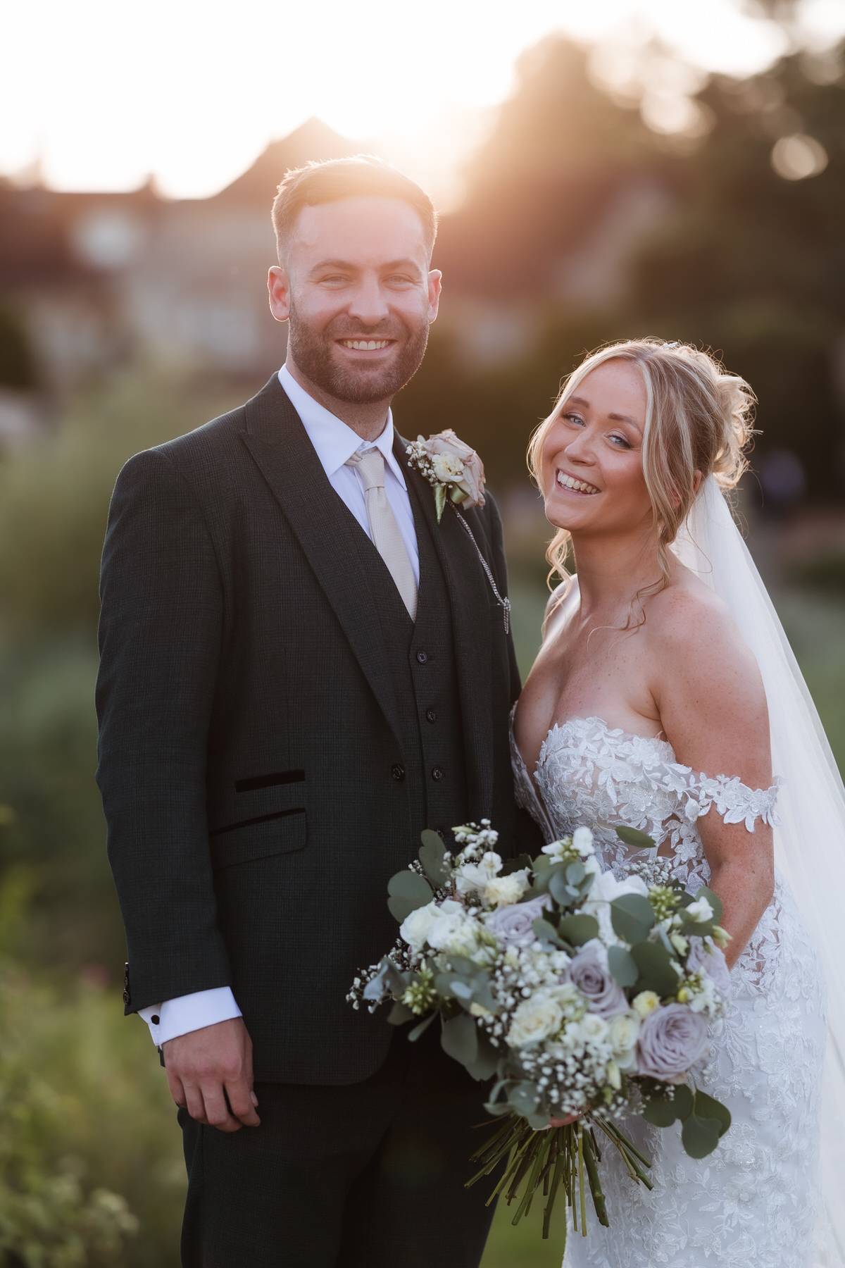 A bride in an off-shoulder lace dress holding a bouquet stands smiling beside a groom in a dark suit and tie. They are outdoors at sunset, with soft sunlight and greenery in the background.