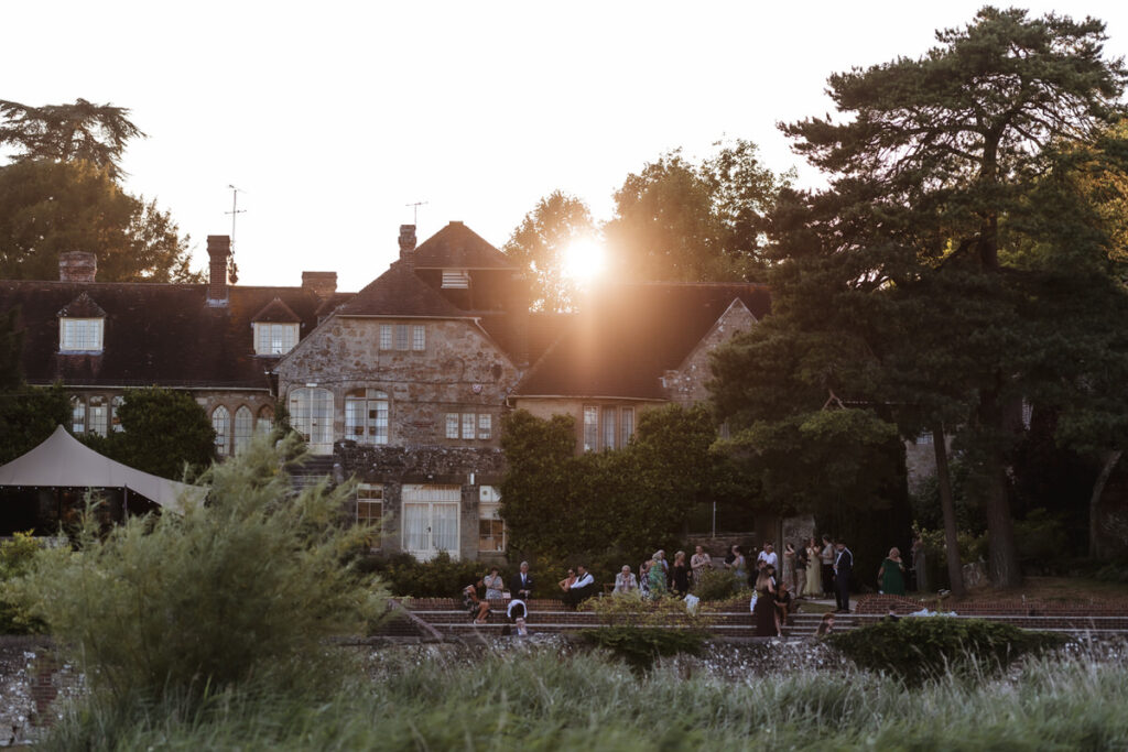 A large stone manor house with ivy-covered walls stands at sunset. People are gathered on the lawn in elegant attire, and tall trees frame the scene, with warm sunlight streaming through the branches.