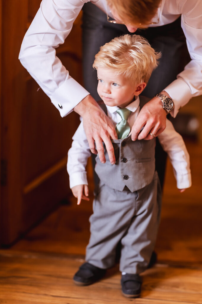 A young boy with blond hair, dressed in a gray suit, vest, and tie, stands still while an adult adjusts his clothing, both on a wooden floor inside a warmly lit room.