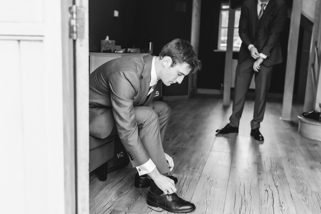 A man in a suit bends down to tie his shoe in a well-lit room with wooden floors, while another man in a suit adjusts his cuff in the background.