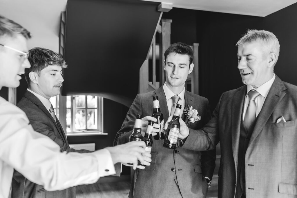 Four men in suits clink beer bottles together and smile indoors, appearing to celebrate. The setting is stylish and the mood is cheerful and relaxed. The photo is in black and white.
