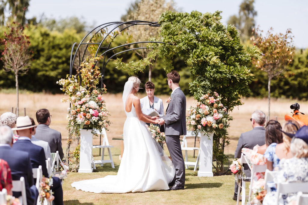 A bride and groom stand facing each other during an outdoor wedding ceremony, holding hands in front of a floral arch, with a celebrant and seated guests watching. The setting is sunny with greenery in the background.