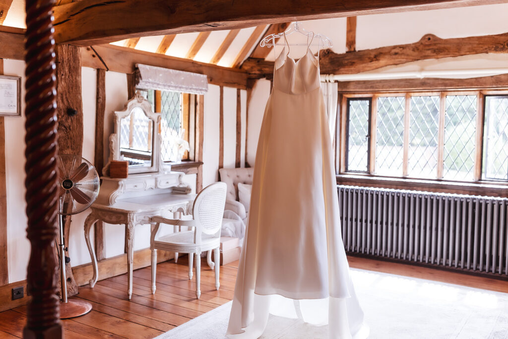 A white wedding dress hangs in a rustic, sunlit room with exposed wooden beams, a vintage white vanity with a mirror, a cushioned chair, and large windows with diamond-patterned glass.