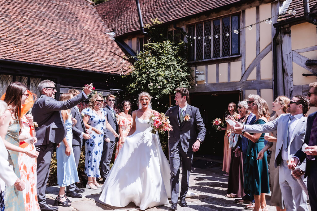 A newlywed couple walks outside, smiling, as guests cheer and throw flower petals. The group is gathered between timbered buildings under sunny skies, dressed in colorful formal attire.