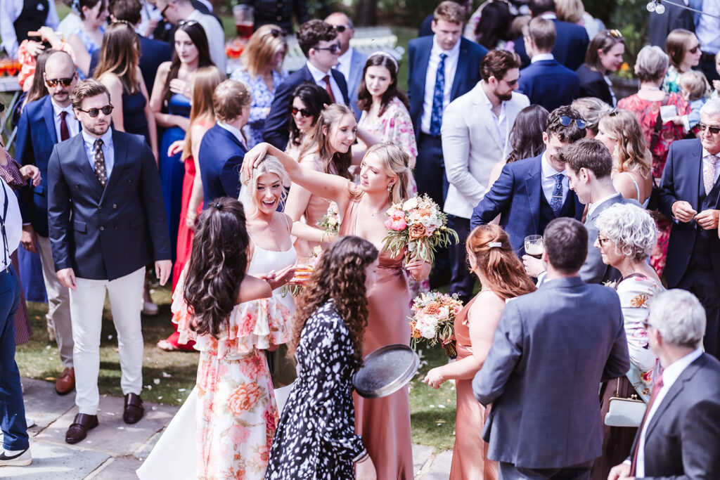 A lively outdoor wedding reception with guests in formal attire mingling. Bridesmaids in pink dresses hold bouquets, and everyone appears to be enjoying the celebration under the sunlight.