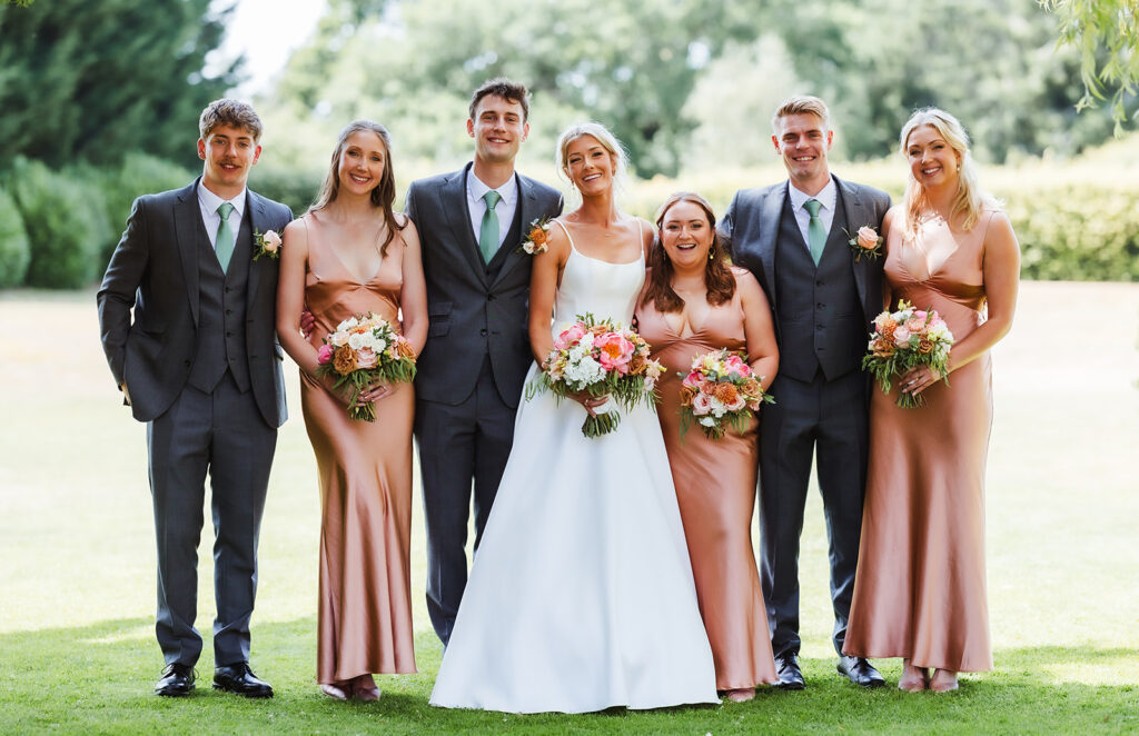 A bride in a white dress stands with her groom and bridal party outdoors. The bridesmaids wear blush pink dresses and hold bouquets, while the groomsmen wear grey suits with green ties. Everyone is smiling.