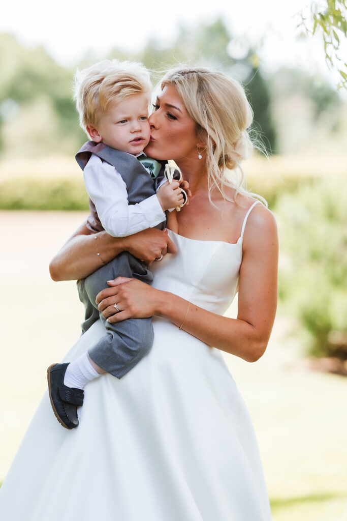 A woman in a white dress holds a young boy in a gray vest and pants, kissing him on the cheek, outdoors on a sunny day with greenery in the background.