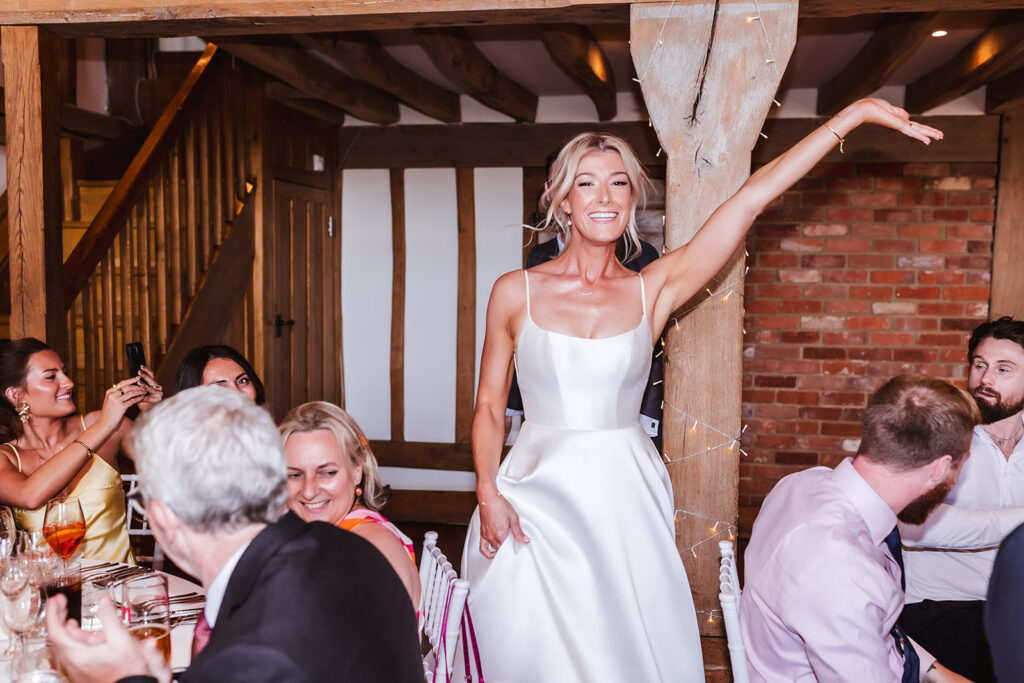 A smiling woman in a white dress stands and raises her arm joyfully at a dining table, surrounded by seated guests in a rustic venue with wooden beams and brick walls.