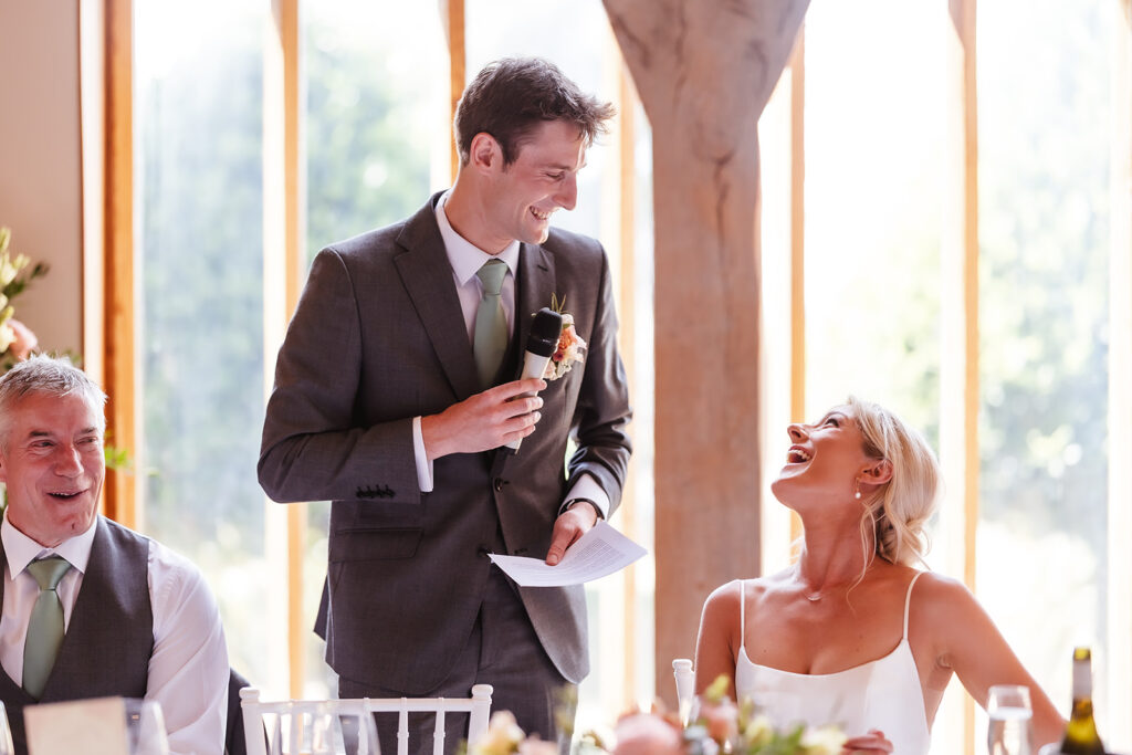 A man in a suit stands holding a microphone and paper, smiling at a seated woman in a white dress who is laughing. Another man sits beside them, also smiling. They are at a bright, elegant indoor event.