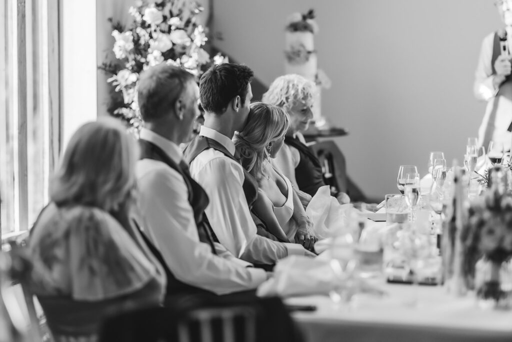 A black and white photo of a wedding reception shows a bride and groom sitting closely together at a table, surrounded by guests, with floral decorations and wine glasses visible.