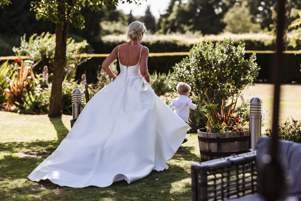 A bride in a white dress walks outside on a sunny day, holding hands with a young child. They are surrounded by greenery and garden plants. The photo is taken from behind.