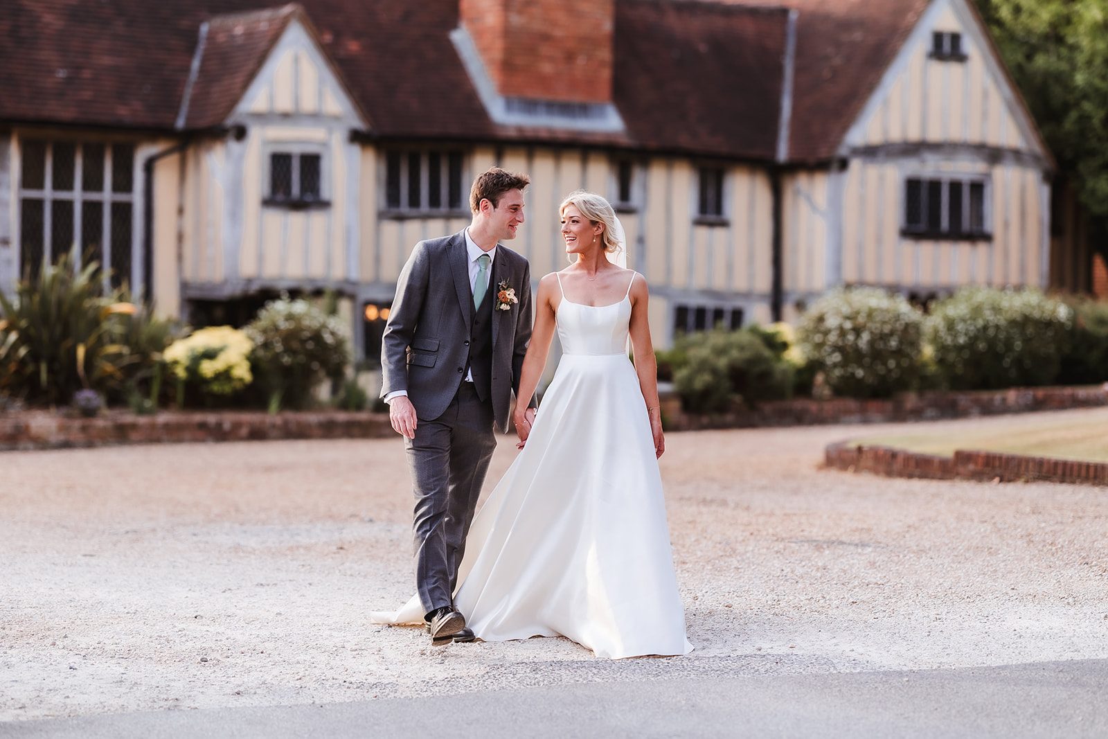 A bride in a white dress and a groom in a gray suit walk hand in hand outside a historic, timber-framed building with a red roof and lush greenery in the background.